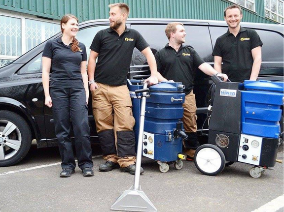 This is a photo of Borehamwood Carpet Cleaning carpet cleaners (three men and one woman) standing in fromt of their black van, with two steam cleaning carpet machines next to them